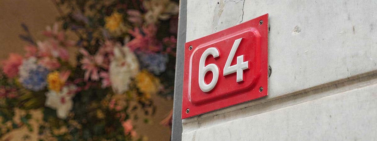 Placa de número de casa roja con el número 64 sobre fachada de mármol blanco con flores coloridas de fondo